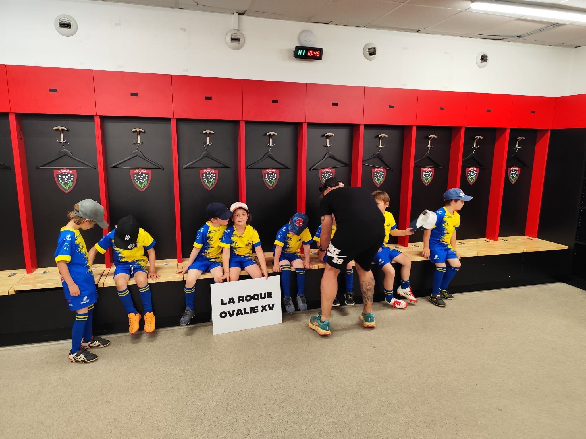 Les jeunes de l'école de rugby dans un vestiaire de stade, derrière la pancarte La Roque Ovalie XV